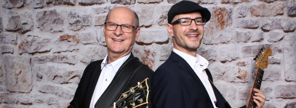 Zwei Männer, lächelnd, in Anzug und Brille, stehen Rücken an Rücken mit Gitarren vor einer strukturierten Steinwand. Einer hält eine elektrische Gitarre, der andere eine akustische Gitarre – ein harmonisches Duo, das perfekt dazu geeignet ist, bei jeder Hochzeit den Ton anzugeben.