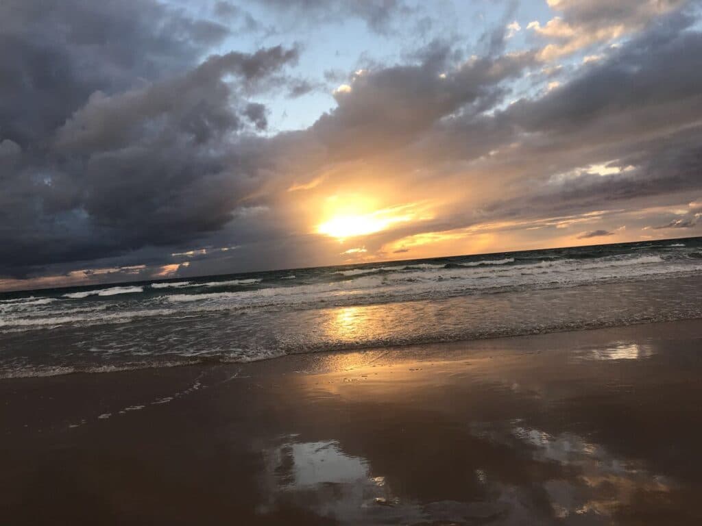 Wenn die Sonne über dem Strand untergeht und ein goldenes Licht auf die Meereswellen und den nassen Sand wirft, umhüllt ein Gefühl der Ruhe die Szene – perfekt für Paare, die von einer malerischen Hochzeit träumen. Der teilweise bewölkte Himmel trägt zu dieser unvergesslichen Kulisse für Ihre Hochzeitsplanung bei.