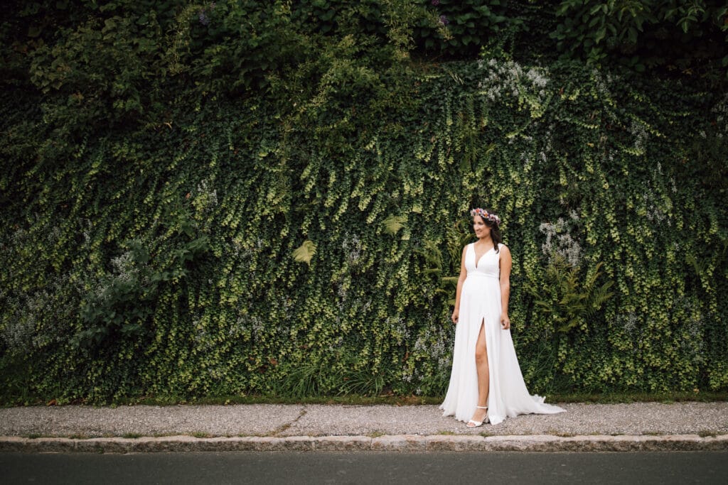 Eine Frau in einem weißen Kleid und einem Blumenkranz steht auf einem Gehweg neben einer üppigen grünen, mit Efeu bewachsenen Mauer und verkörpert die ruhige Atmosphäre einer Traumhochzeit. Ihr Kleid mit dem hohen Schlitz fängt die Essenz zeitloser Eleganz ein.