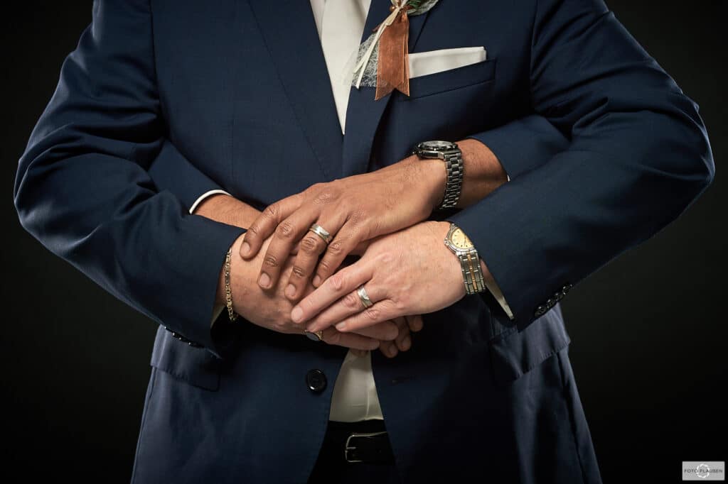 Zwei Menschen in Anzügen umarmen sich, wobei nur ihre Oberkörper und gefalteten Hände vor einem dunklen Hintergrund zu sehen sind. Beide tragen Uhren und Ringe, wobei an einem Anzug eine Boutonniere befestigt ist – ein Symbol der Intimität, das oft in der Hochzeitsplanung zu sehen ist und ihre enge Bindung unterstreicht.