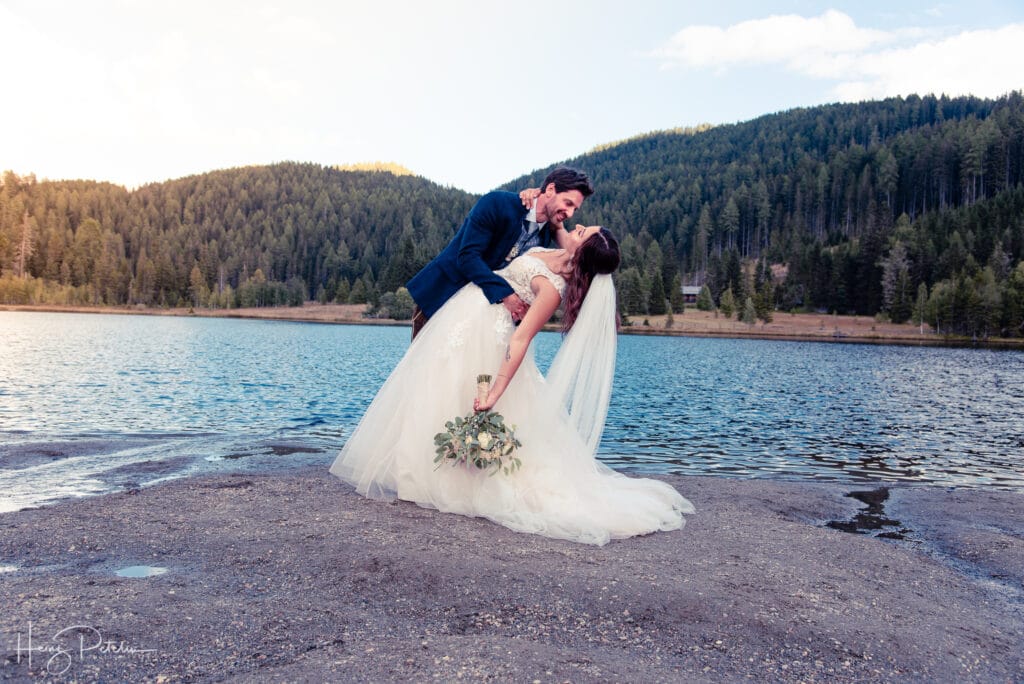 Ein Bräutigam im dunklen Anzug taucht die Braut, die ein fließendes weißes Kleid und einen Schleier trägt, sanft ins Wasser, während sie sich während ihrer Hochzeit am ruhigen See küssen. Im Hintergrund sind ein üppiger grüner Wald und ein klarer Himmel zu sehen, was die romantische Atmosphäre dieses perfekten Moments auf ihrer Hochzeitsreise noch verstärkt.