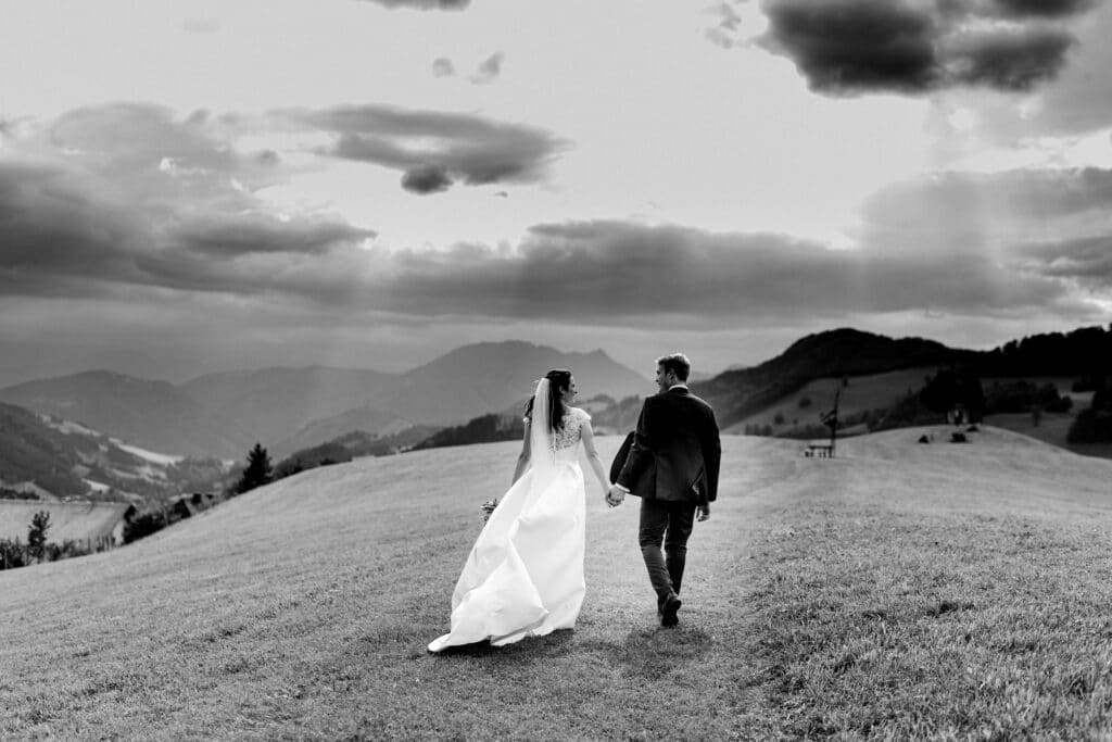 Ein Schwarzweißfoto fängt einen Hochzeitsmoment ein, als Braut und Bräutigam Hand in Hand über eine grasbewachsene Hügelkuppe gehen. Das Kleid der Braut weht anmutig hinter ihr her, vor der Kulisse sanfter Hügel und dramatischer Wolken am Himmel.