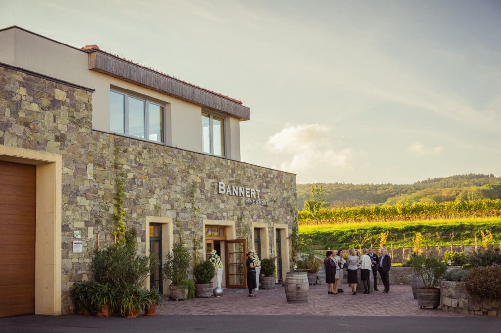 Ein Weingut aus Stein mit einem „Banneret“ an der Fassade ist von Grün umgeben und perfekt für eine traumhafte Hochzeit. Mehrere Personen stehen draußen neben Holzfässern unter einem teilweise bewölkten Himmel. In der Ferne erstrecken sich sanfte Weinberge und bilden eine ideale Kulisse für die Hochzeitsplanung.