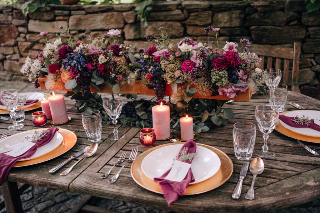 Ein rustikaler Esstisch im Freien ist mit eleganten Tellern, Gläsern und Besteck gedeckt. Ein Tafelaufsatz aus leuchtenden Blumen und Trauben schmückt den Tisch neben brennenden Kerzen. Servietten mit Hochzeitsmotiven verleihen vor der Steinmauer im Hintergrund eine persönliche Note.