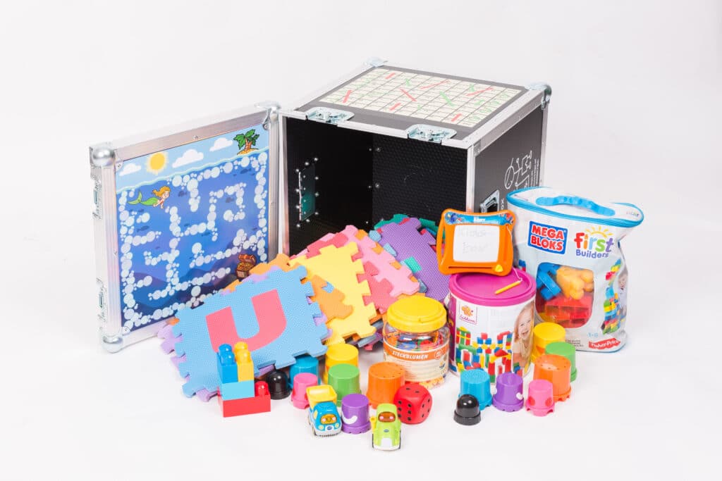 Eine Sammlung farbenfroher Spielzeuge und Spiele, darunter Puzzleteile, Mega Bloks und Lernwürfel, umgibt eine schwarze Aufbewahrungsbox – ideal, um die Kleinen zu unterhalten, während Sie sich auf die Hochzeitsplanung konzentrieren. Ein kleines magnetisches Zeichenbrett und ein Aktivitätspanel mit Labyrinthen sind ebenfalls sichtbar.