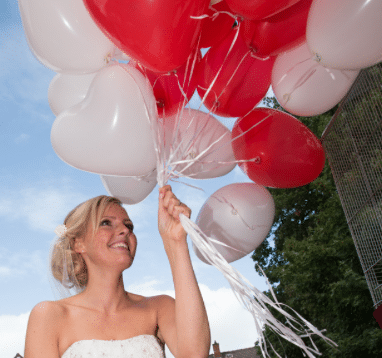 Eine lächelnde Person in einem weißen Kleid hält einen großen Strauß roter und weißer herzförmiger Luftballons vor einem teilweise bewölkten Himmel und fängt die Essenz der Hochzeitsplanung an ihrem besonderen Tag ein.
