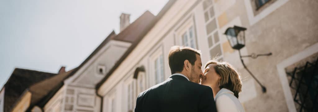 Ein Paar küsst sich vor charmanten, altmodischen Gebäuden unter einem strahlenden Himmel und stellt sich seine Hochzeit vor. Die hellen Gebäude sind mit dekorativen Elementen ausgestattet und eine Vintage-Laterne schmückt die Wand daneben – eine perfekte Kulisse für ihre Hochzeitsplanung.