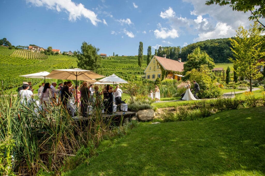Eine malerische Hochzeit im Freien mit Dutzenden von Gästen, die sich unter Sonnenschirmen an einem Teich versammelt haben. Üppiges Grün umgibt das Gebiet, mit Weinbergen und einem rustikalen Haus im Hintergrund. Braut und Bräutigam stehen zusammen auf einem Weg und schaffen eine romantische Atmosphäre, die perfekt für die Hochzeitsplanung ist.