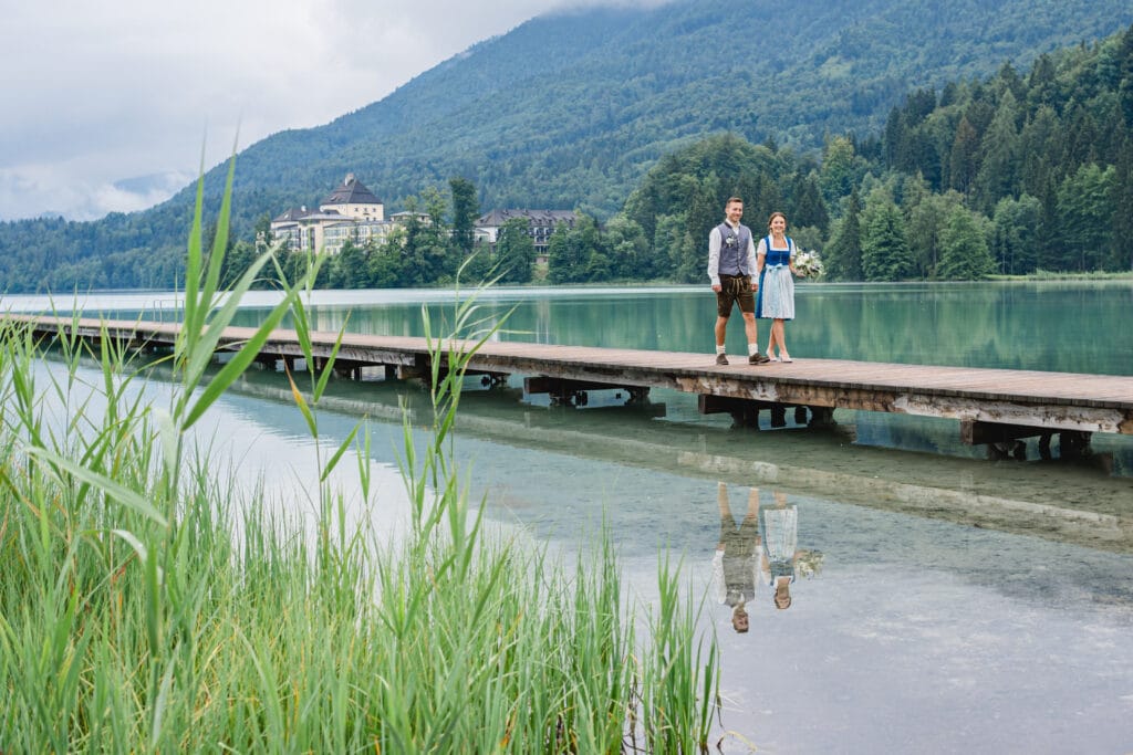 Ein Paar in traditioneller Kleidung steht auf einem Holzsteg über einem klaren See, umgeben von üppigem Grün und Bergen. Ihr Spiegelbild schimmert im Wasser und fängt die Essenz ihrer Hochzeit ein. Ein entferntes Gebäude ziert die Hügel und verleiht dieser malerischen Kulisse Charme.