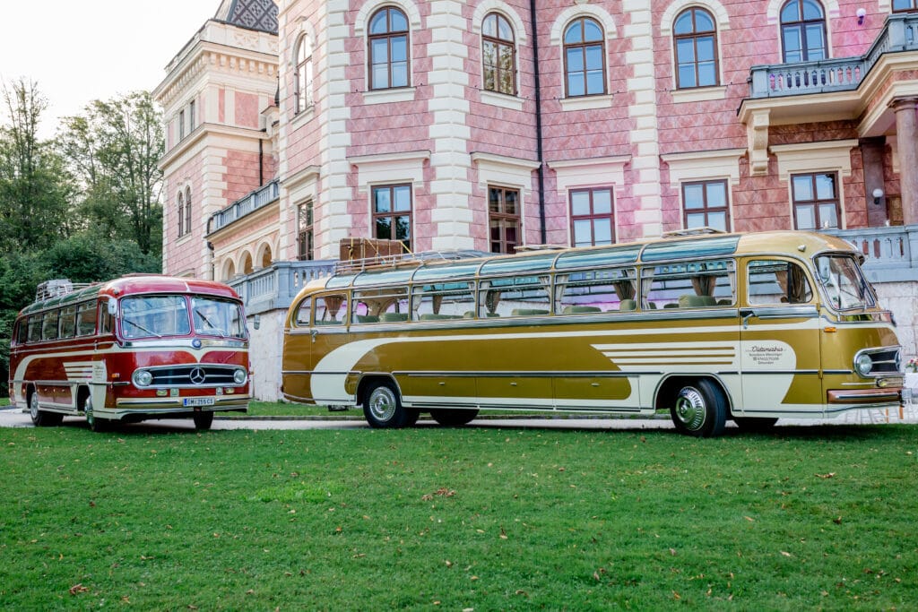 Zwei Oldtimerbusse, ideal für eine Hochzeit, stehen auf einer Rasenfläche vor einem historischen Gebäude aus Rosa und Stein mit Bogenfenstern. Die klassischen Designs in Rot und Gelb mit weißen Details bieten die perfekte Kulisse, während Bäume die malerische Szene sanft einrahmen.