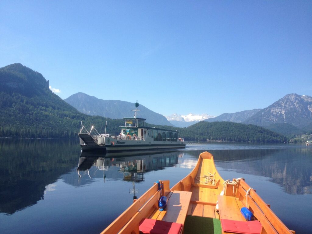 Im Vordergrund ist ein Holzboot mit bunten Sitzen zu sehen, das auf einem ruhigen See schwimmt und sich perfekt für eine Hochzeitskulisse eignet. In der Ferne gleitet eine Fähre vorbei. Die umliegenden, mit Kiefern bewachsenen Berge und der klare, blaue Himmel spiegeln sich wunderschön im Wasser und bilden eine ideale Kulisse für die Hochzeitsplanung.