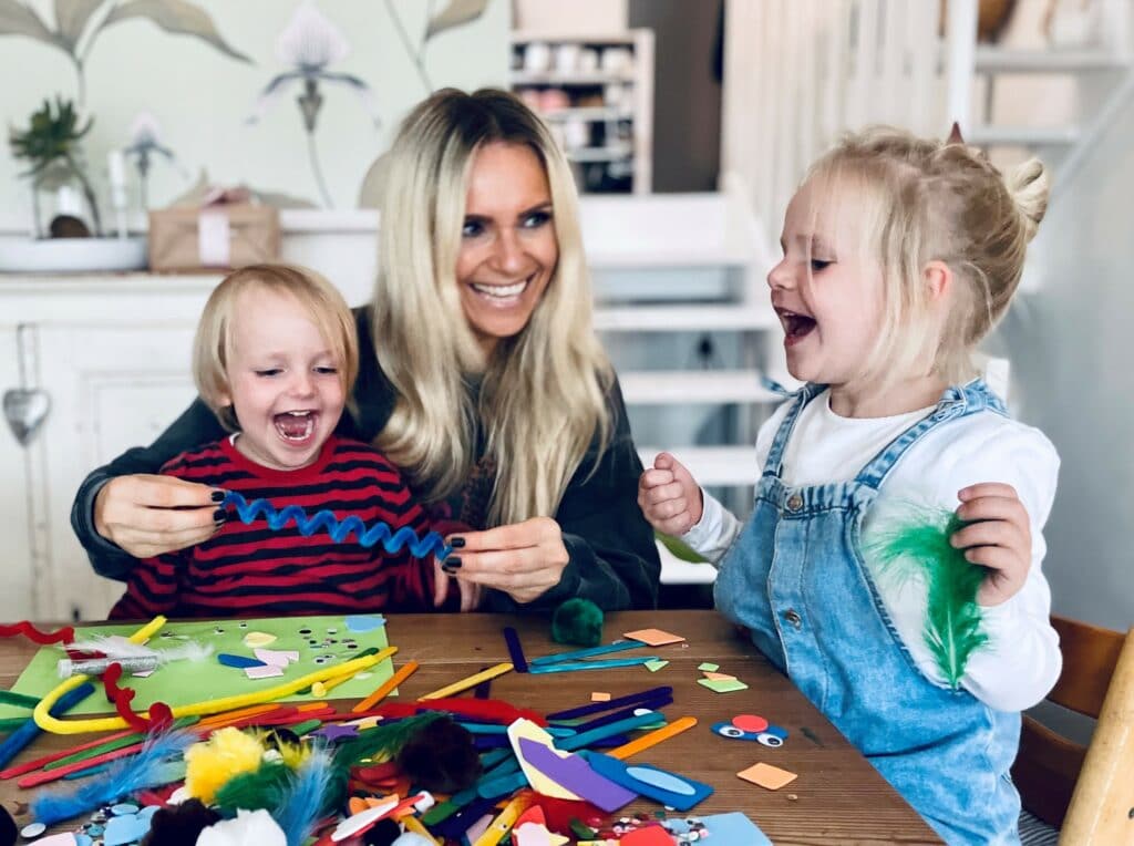 Eine Frau und zwei Kinder lächeln und basteln an einem Tisch voller bunter Materialien wie Pfeifenreiniger, Schaumstofffiguren und Federn, vielleicht um Dekorationen für eine fröhliche Hochzeit zu basteln. Der Raum hat eine fröhliche Atmosphäre mit hellem Dekor im Hintergrund.