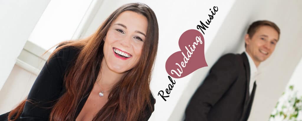 Eine lächelnde Frau mit langen braunen Haaren und ein Mann im Anzug stehen vor einer weißen Wand. Die Worte „Real Wedding Music“ sind elegant in Herzform daneben angebracht, perfekt für jede Hochzeits- oder Heiratsfeier.