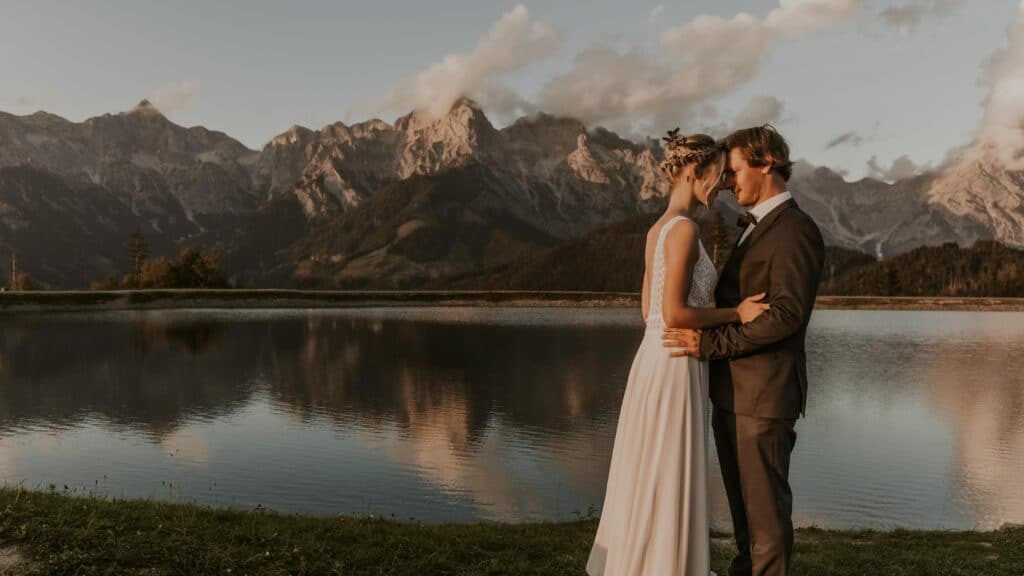 Ein Paar in Hochzeitskleidung umarmt sich an einem ruhigen See, auf dessen Oberfläche sich schneebedeckte Berge und ein teilweise bewölkter Himmel spiegeln. Diese idyllische Szene fängt die Romantik ihrer Hochzeit ein und zeigt einen friedlichen Moment in der Natur.