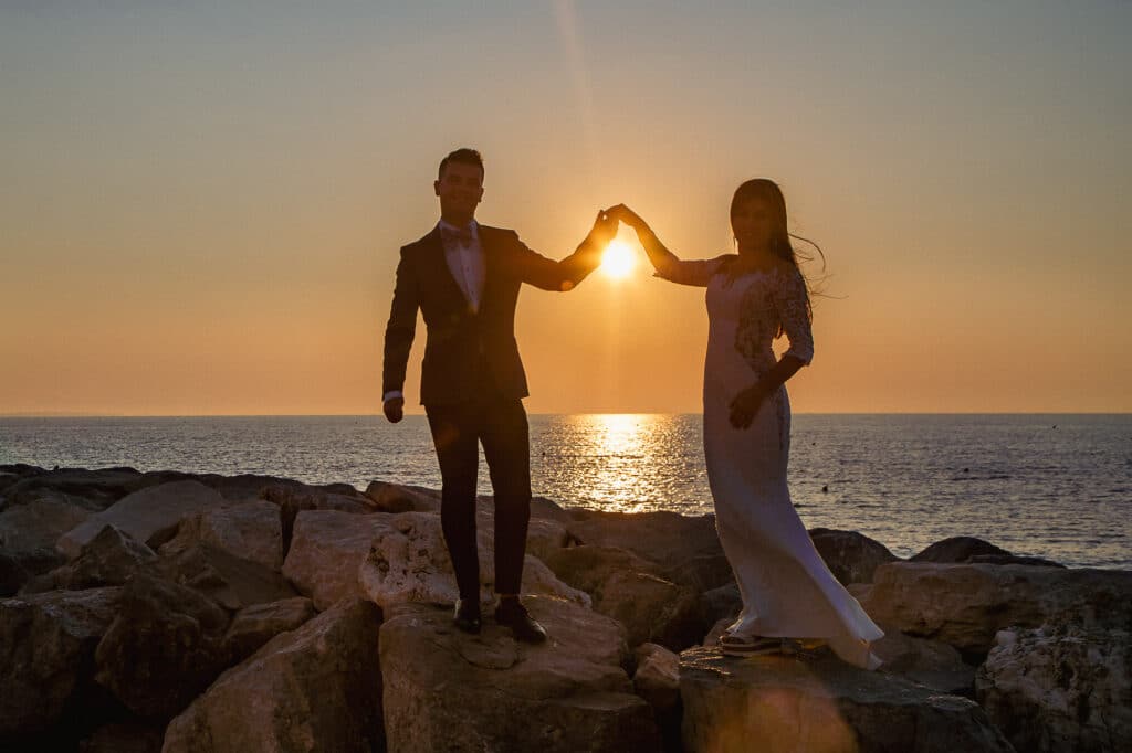 Ein Paar steht bei Sonnenuntergang auf Felsen am Meer, hält Händchen, ihre Herzen sind eingehüllt in die Magie der Hochzeit. Die Sonne geht zwischen ihnen unter und verbreitet einen warmen Schein. Die Frau trägt ein weißes Kleid und der Mann einen Anzug. Der Himmel ist in Orange und Blau getaucht, perfekt für ihre verträumte Hochzeitsplanung.