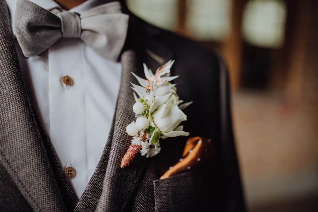 Nahaufnahme eines Bräutigams in dunkelgrauem Anzug und Fliege, bereit für die Hochzeit. Eine weiße Ansteckblume mit kleinen Blumen und Grün ziert das Revers, während ein senffarbenes Einstecktuch aus der Jackentasche hervorschaut, perfekt gestylt für diesen besonderen Tag.