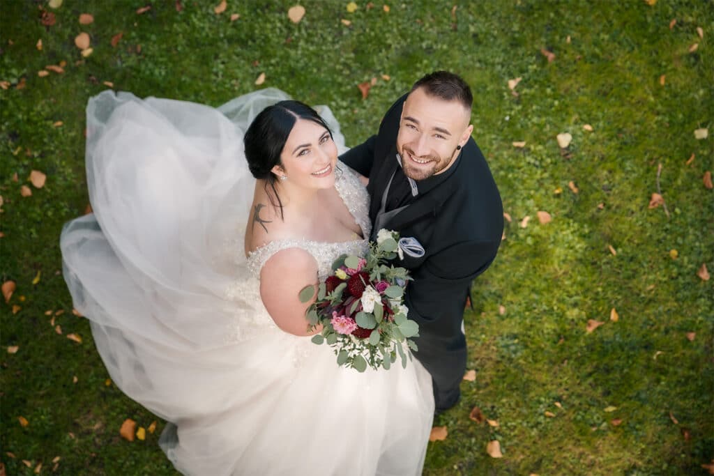 Braut und Bräutigam lächeln an ihrem Hochzeitstag in die Kamera. Die Braut in einem weißen Brautkleid hält einen Strauß roter und rosa Blumen, während der Bräutigam einen dunklen Anzug trägt. Sie stehen auf grünem Gras mit verstreuten Blättern und fangen ihren perfekten Moment des Eheglücks ein.