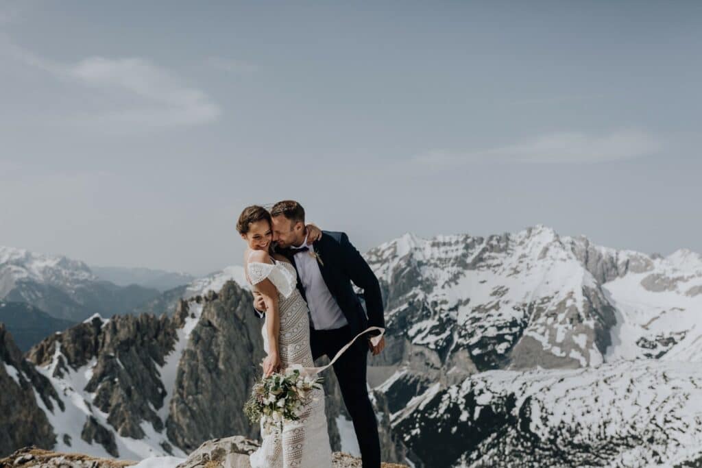 Ein Paar steht auf einem Berggipfel mit schneebedeckten Gipfeln im Hintergrund und fängt einen perfekten Hochzeitsmoment ein. Der Mann im Anzug küsst die Frau liebevoll auf die Wange. Sie trägt ein weißes Spitzenkleid und hält einen Blumenstrauß in der Hand. Beide lächeln inmitten der atemberaubenden Landschaft.