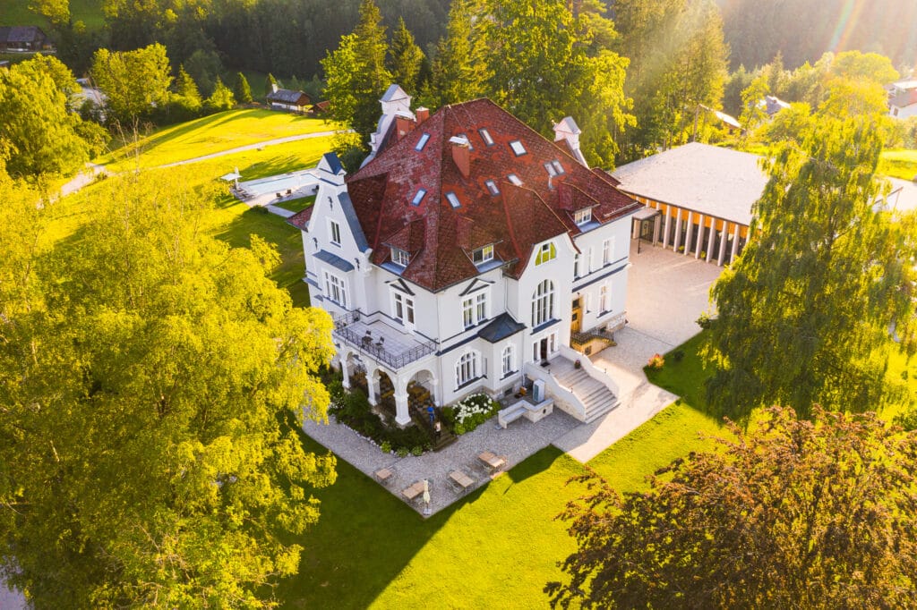 Luftaufnahme eines großen, eleganten Herrenhauses, das sich ideal für die Hochzeitsplanung eignet und von üppigem Grün umgeben ist. Das Gebäude verfügt über ein rotes Satteldach mit mehreren Dachgauben, weißen Wänden und Bogenfenstern. Sonnenlicht fällt durch die umliegenden Bäume und schafft eine ruhige Atmosphäre, die perfekt für eine Traumhochzeit ist.