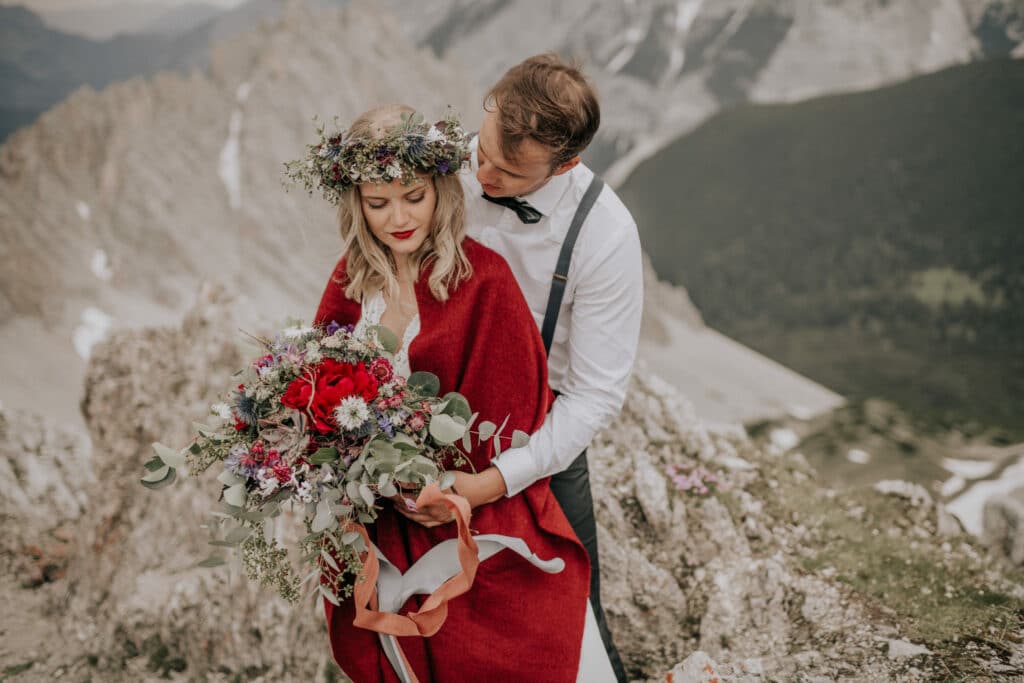 Ein Paar umarmt sich in einer felsigen Berglandschaft und feiert seine Hochzeit. Die Frau, in einen roten Schal gehüllt und mit einer Blumenkrone bekleidet, hält einen farbenfrohen Blumenstrauß in der Hand. Der Mann steht in einem weißen Hemd und Hosenträgern dahinter. Schneebedeckte Gipfel bilden die atemberaubende Kulisse für ihren besonderen Moment.