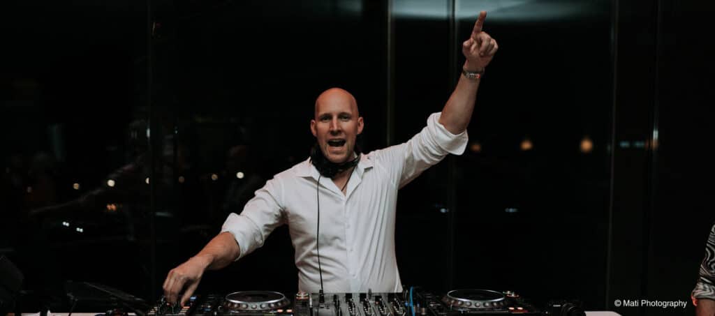 Ein kahlköpfiger Mann in weißem Hemd steht in einem schwach beleuchteten Raum hinter einer DJ-Anlage. Er trägt Kopfhörer um den Hals und hebt einen Arm nach oben, während er spricht oder singt. Damit verwandelt er die Atmosphäre in eine elektrisierende Feier, die perfekt zu jeder Hochzeit passt.