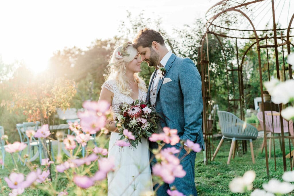 Braut und Bräutigam stehen eng beieinander in einem sonnenbeschienenen Garten und verkörpern damit die Essenz einer bezaubernden Hochzeit. Die Braut hält einen Blumenstrauß und trägt eine Blumenkrone, während der Bräutigam einen blauen Anzug trägt. Im Vordergrund blühen rosa Blumen, und bezaubernde Gartenstrukturen runden diese ruhige Szene ab.