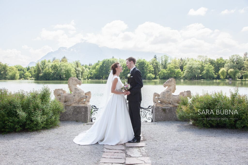 Braut und Bräutigam stehen sich gegenüber und halten sich an den Händen vor einem ruhigen See. Dies verkörpert die Essenz einer perfekten Hochzeit. Sie sind von üppigem Grün und steinernen Pferdeskulpturen umgeben, im Hintergrund erhebt sich unter einem teilweise bewölkten Himmel ein majestätischer Berg.