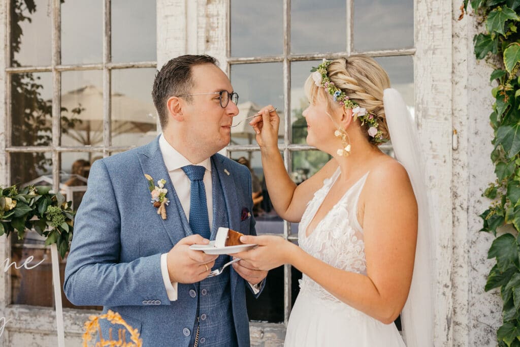 Das Paar feiert freudig seine Hochzeit. Der Bräutigam im blauen Anzug hält einen Teller mit Kuchen, während die Braut in ihrem weißen Kleid ihm spielerisch einen Bissen gibt. Sie stehen an einem rustikalen Fenster mit üppigem Grün und halten den perfekten Moment ihrer Hochzeit fest.