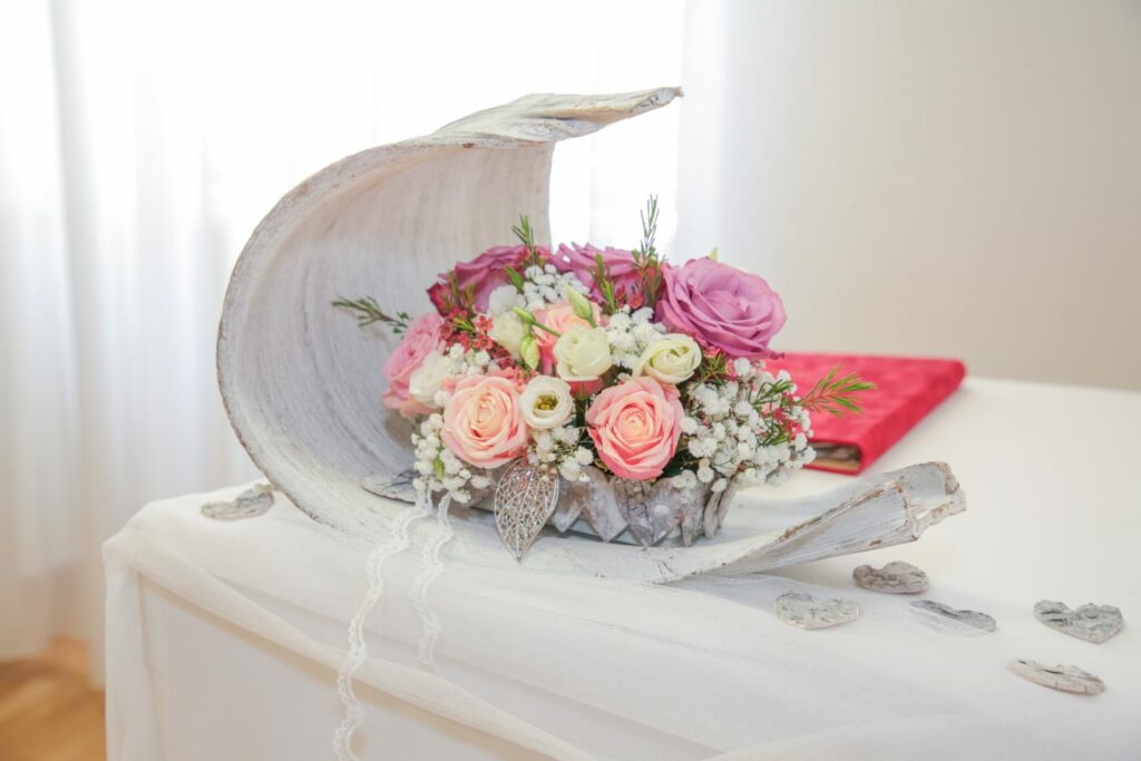 Ein dekoratives Arrangement mit rosa und weißen Rosen, Schleierkraut und grünen Zweigen in einem rustikalen, halbmondförmigen Holzbehälter ist perfekt für Ihre Hochzeit. Es steht auf einem weiß gedeckten Tisch neben herzförmigen Ornamenten und wertet jedes Hochzeitsdekor auf.