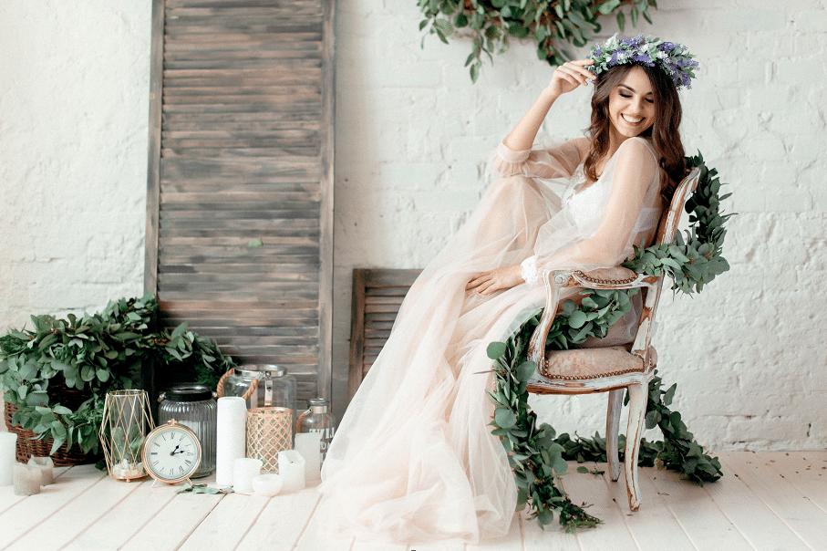Eine Frau in einem durchsichtigen weißen Kleid und mit Blumenkrone sitzt auf einem mit Grünpflanzen geschmückten Stuhl und strahlt Freude aus, während sie die Hochzeit plant. Umgeben von Kerzen, einer Uhr und Blattdekorationen vor einer weißen Backsteinwand ist dies eine zauberhafte Kulisse für ihre Heiratsträume.
