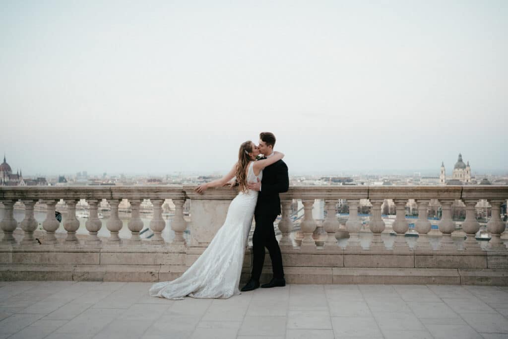Ein Paar in Hochzeitskleidung umarmt sich auf einer Steinterrasse mit einer Stadtlandschaft im Hintergrund und symbolisiert so ihre Reise in die „Hochzeit“. Das lange Kleid der Braut weht elegant, während der Bräutigam in einem dunklen Anzug mit ihr über die ferne Skyline unter einem blassblauen Himmel blickt.