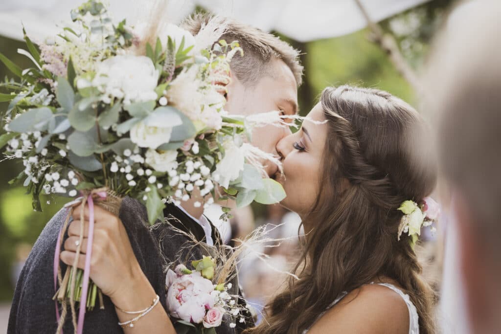Ein Paar küsst sich an seinem Hochzeitstag, versunken in die Romantik des Heiratens. Die Braut hält einen großen Blumenstrauß mit Grünpflanzen, während der Bräutigam sie umarmt. Im Freien, umgeben von verschwommenem Grün, fängt die Szene die freudige Essenz sorgfältiger Hochzeitsplanung ein.