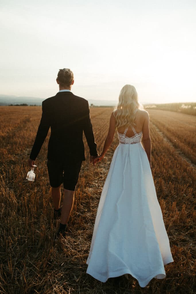 Ein Paar geht Händchen haltend durch ein sonnenbeschienenes Feld und versunken in die Schönheit seiner Hochzeit. Die Frau trägt ein fließendes weißes Kleid und der Mann ist im Anzug und trägt eine kleine Glasflasche. Während die Sonne am Horizont untergeht, wirft sie einen warmen Schein auf diesen magischen Moment der Liebe.