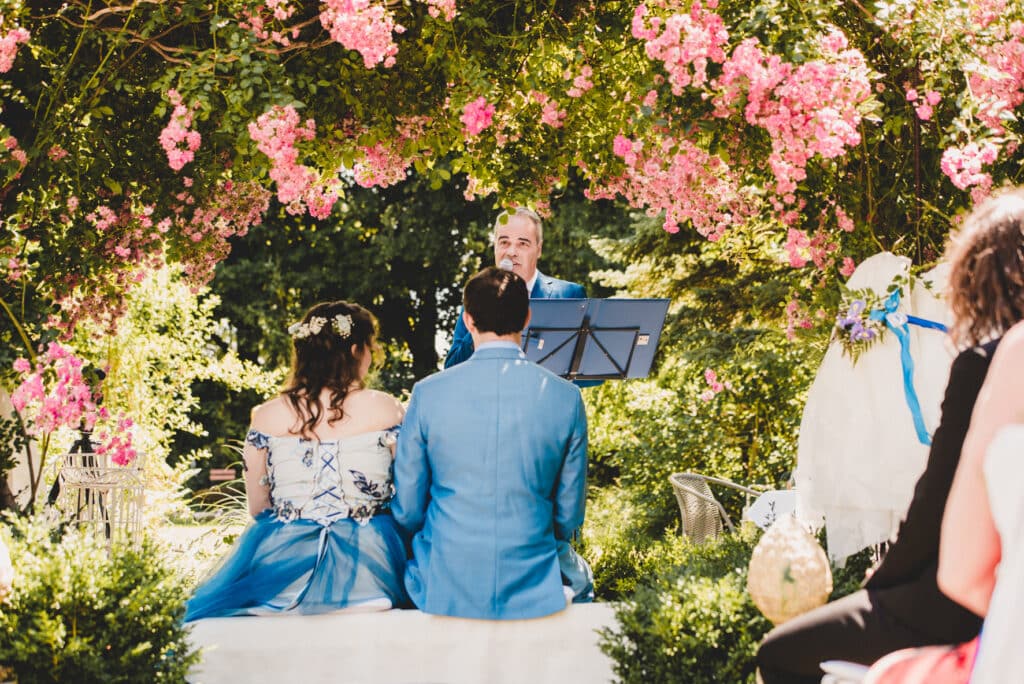 Ein Paar sitzt während seiner Hochzeit auf einer weißen Bank und blickt einem Geistlichen gegenüber. Die Zeremonie findet im Freien statt, umrahmt von leuchtend rosa Blumen und üppigem Grün, während der Geistliche hinter einem Podium unter einem Blumenbogen steht – eine idyllische Szene, wie aus den Träumen der Hochzeitsplanung.