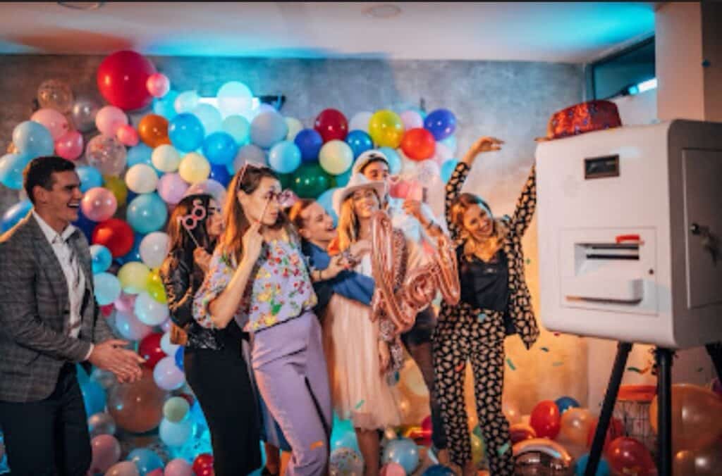 Eine Gruppe von Menschen posiert fröhlich vor einer Fotokabine bei einer farbenfrohen Hochzeitsfeier. Der Hintergrund ist mit zahlreichen Luftballons in verschiedenen Farben dekoriert. Eine Person hält einen Hut, während andere verspielte Posen einnehmen und so eine festliche Atmosphäre schaffen, die perfekt für Hochzeitserinnerungen ist.