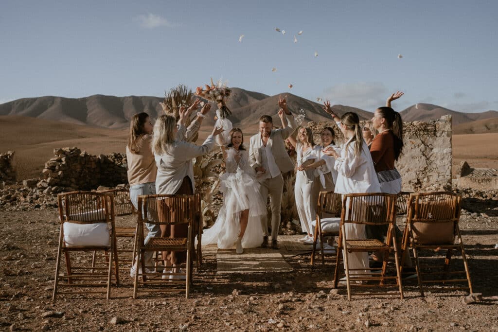 Ein frisch vermähltes Paar schreitet durch einen rustikalen Gang im Freien, umgeben von fröhlichen Gästen, die Blütenblätter werfen. Vor der Kulisse trockener Berge unter einem klaren blauen Himmel findet ihre Hochzeit mit Holzstühlen im Vordergrund statt. Die Szene ist festlich und feierlich.