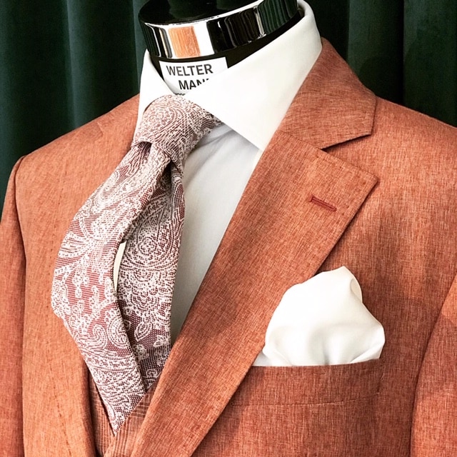 Eine Nahaufnahme einer Schaufensterpuppe präsentiert einen strukturierten rostfarbenen Anzug, perfekt für diejenigen, die ihre Hochzeit planen, kombiniert mit einem weißen Hemd und einer Krawatte mit Paisleymuster. Ein weißes Einstecktuch steckt ordentlich in der Brusttasche des Anzugs.