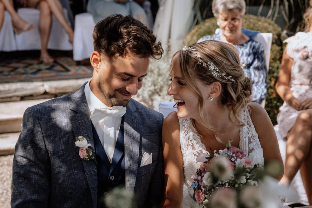 Ein lächelndes Paar sitzt während einer Hochzeitszeremonie im Freien eng beieinander. Die Braut hält einen Blumenstrauß in der Hand und trägt ein Spitzenkleid und einen Blumenkopfschmuck, was die Essenz des Hochzeitsglücks verkörpert. Der Bräutigam trägt einen karierten Anzug mit Fliege. Im Hintergrund sind einige entspannte Gäste zu sehen.