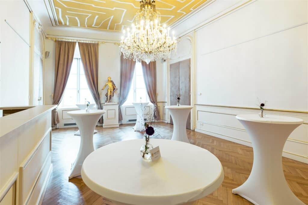 Ein eleganter Raum mit hohen Decken, perfekt für eine Hochzeit, verfügt über vier runde Cocktail-Tische, die mit weißen Tüchern behangen sind. Ein großer Kronleuchter hängt von der verzierten Decke, während eine goldene Statue stolz neben großen Fenstern steht, die mit üppigen Vorhängen geschmückt sind.