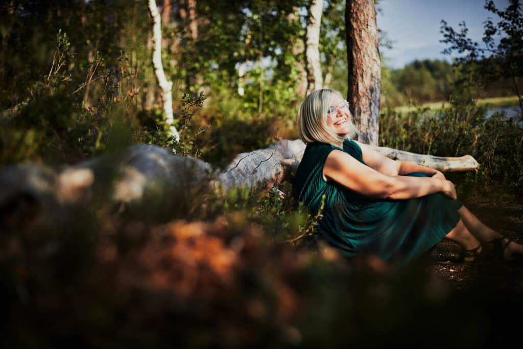 Eine Frau mit grauem Haar und Brille sitzt lächelnd vor einem Baum im Wald und erinnert an die ruhigen Momente, die man bei der Hochzeitsplanung oft erlebt. Sie trägt ein dunkelgrünes Kleid, das Sonnenlicht fällt durch die Blätter und wirft gesprenkelte Schatten auf den Boden. Die Szene vermittelt Ruhe und Natur.