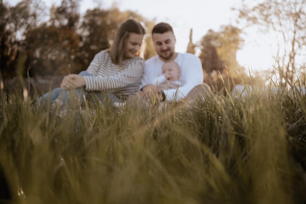 Eine dreiköpfige Familie sitzt zusammen auf einer Wiese. Die Eltern, leger gekleidet und lächelnd, blicken liebevoll auf ihr Baby. Während die Sonne untergeht und einen warmen Schein über die Szene wirft, schwelgen sie in Erinnerungen an ihren Hochzeitstag und träumen davon, zukünftige besondere Momente gemeinsam zu planen.