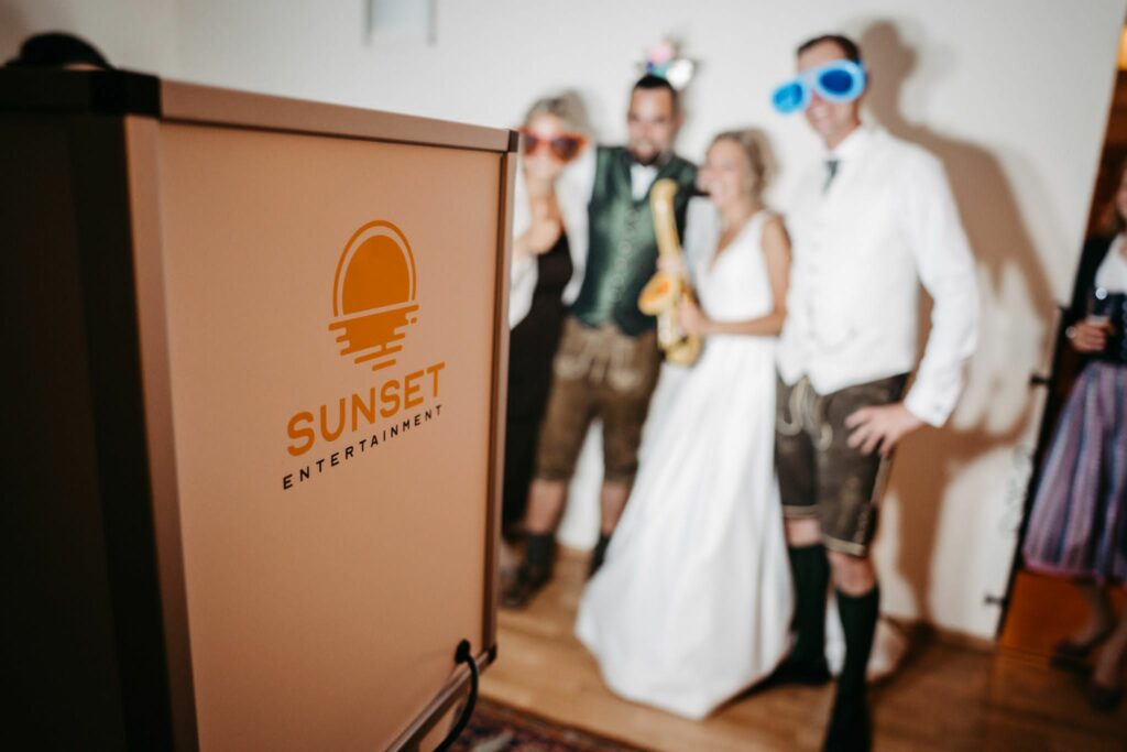 Eine Gruppe von Menschen in festlicher Kleidung, darunter übergroße Sonnenbrillen, posiert für ein Foto in der Nähe eines Standes mit dem Logo von „Sunset Entertainment“. Sie scheinen drinnen etwas zu feiern, das wie eine Hochzeit aussieht, und schaffen eine fröhliche Atmosphäre.