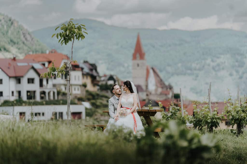 Ein Paar in Hochzeitskleidung sitzt auf einer Picknickbank in einer üppig grünen Landschaft und verkörpert die Romantik ihrer Hochzeit. Hinter ihnen schmiegen sich ein malerisches Dorf und ein Kirchturm an sanfte Hügel und bilden unter dem friedlichen, bewölkten Himmel eine ideale Kulisse für Heiratsträume.