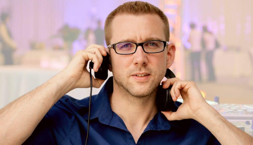 Ein Mann in blauem Hemd und Brille hält sich Kopfhörer vor die Ohren, während die Hochzeitsplanung im Trubel stattfindet. Er steht im Innenbereich, eingerahmt von einem unscharfen Hintergrund aus Menschen und eleganter Hochzeitsdekoration.