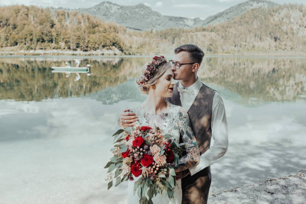 Ein Paar in Hochzeitskleidung steht an einem ruhigen See und verkörpert eine perfekte Hochzeit. Die Braut hält einen leuchtenden Strauß roter und rosa Blumen, während der Bräutigam sie von hinten umarmt. Bäume und Berge spiegeln sich im Wasser unter einem teilweise bewölkten Himmel.