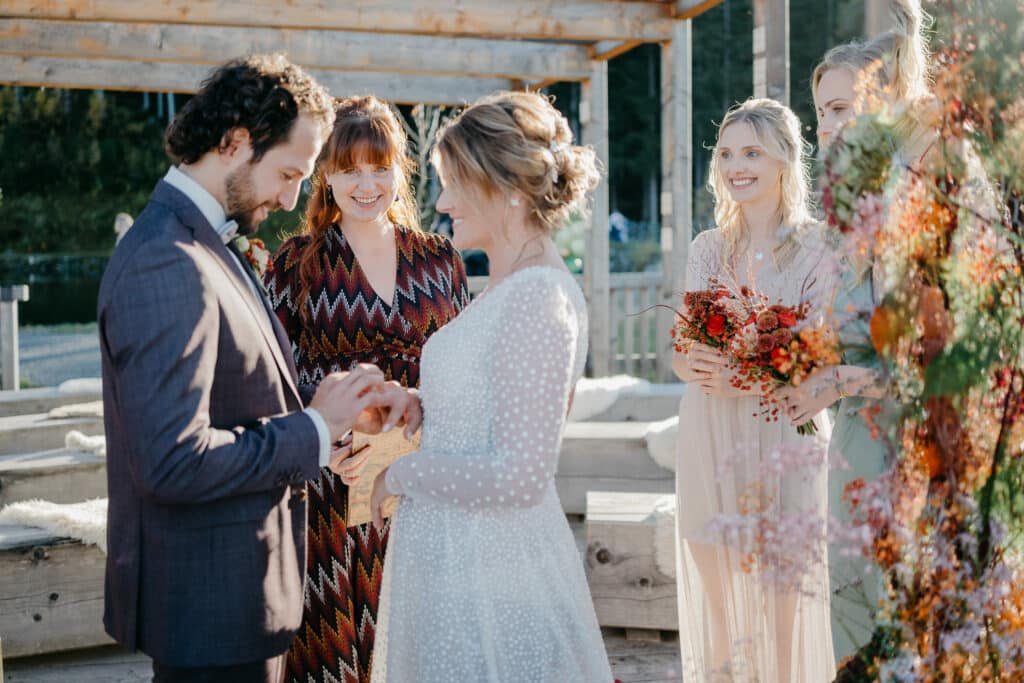 Bei einer bezaubernden Hochzeit im Freien tauschen Braut und Bräutigam die Ringe aus. Die Braut strahlt in ihrem weißen Kleid, während der Bräutigam in seinem Anzug elegant aussieht. Brautjungfern mit eleganten Blumensträußen stehen in der Nähe und strahlen alle freudig in der sonnenbeschienenen Umgebung. Dieser Moment ist perfekt für jeden, der seine eigene Hochzeitsplanung plant.