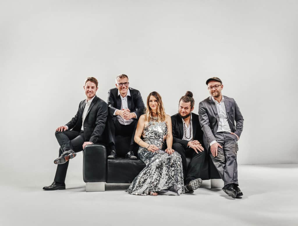 Eine Gruppe von fünf Personen sitzt auf einem schwarzen Sofa vor einem schlichten Hintergrund. Sie sind in formelle Kleidung gekleidet und besprechen vielleicht eine bevorstehende Hochzeit. Die Frau in der Mitte glänzt in einem funkelnden silbernen Kleid, während die Männer Anzüge tragen – einer mit einer Kappe und ein anderer mit einem Dutt – und alle lächeln herzlich.
