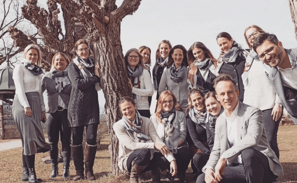 Eine Gruppe von Menschen, bestehend aus Männern und Frauen, posiert im Freien auf einer Rasenfläche. Sie sind ähnlich gekleidet, hauptsächlich in Grau und Weiß mit Kopftüchern. Einige stehen, andere knien, umgeben von Bäumen – eine heitere Szene der Hochzeitsfeierlichkeiten.