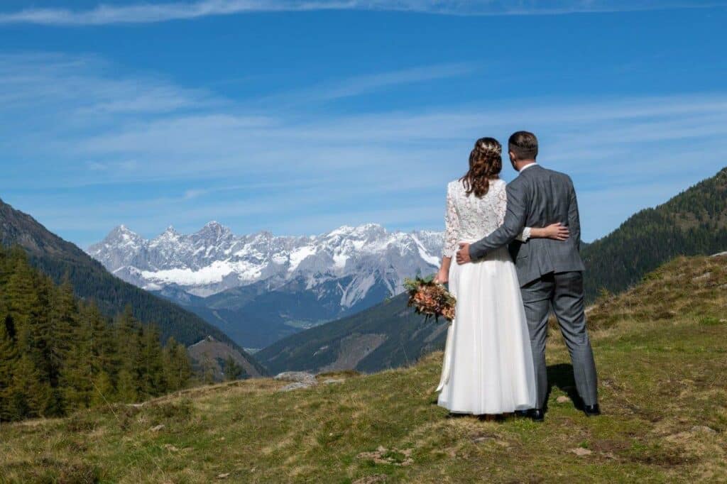 Ein Ehepaar steht auf einem grasbewachsenen Hügel, umgeben von der ruhigen Schönheit einer atemberaubenden Berglandschaft, perfekt für ihre Hochzeit. Die Braut trägt ein weißes Kleid und hält einen Blumenstrauß, und der Bräutigam ergänzt sie in einem grauen Anzug unter dem klaren blauen Himmel.
