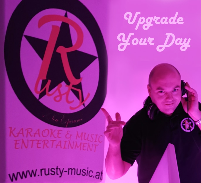 Ein Mann mit Kopfhörern steht neben einem Banner mit der Aufschrift „Rusty Karaoke & Music Entertainment“ und der Website „www.rusty-music.at“. Der rosa beleuchtete Hintergrund flüstert Romantik, perfekt für jede Hochzeit, und „Upgrade Your Day“ oben rechts lädt Freude in Ihre Hochzeitsplanung ein.